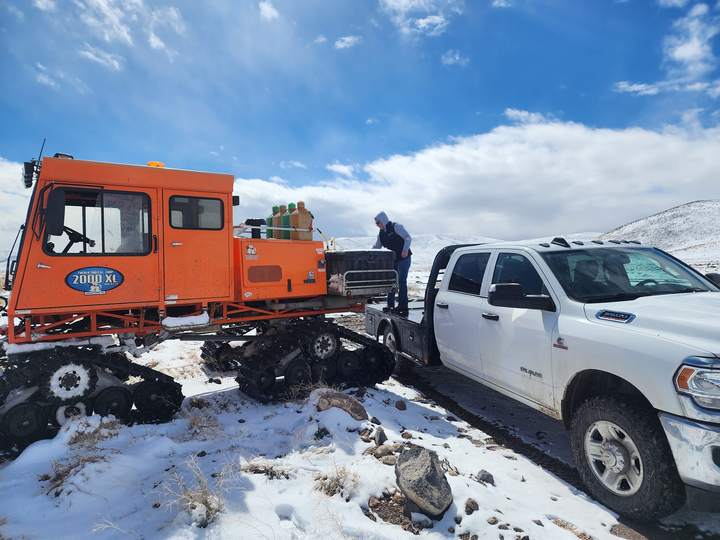 High Desert's orange snowcat beside a white Ram pickup on a snowy mountaintop while a crew member loads fuel cans